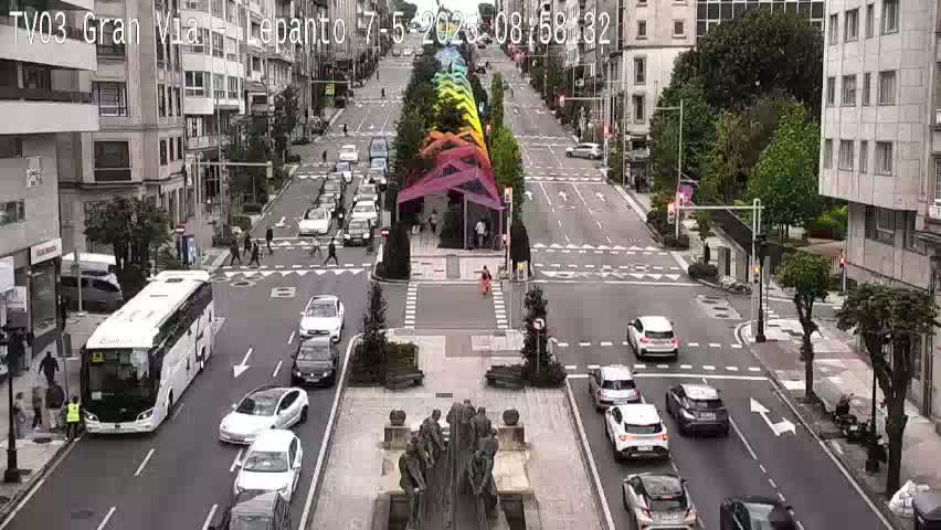 A city street scene on a partly sunny day shows several cars and a bus moving along a wide road that intersects with crosswalks, pedestrians walking, and a colorful structure in the center of the road.