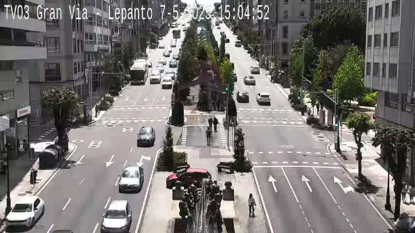 A sunny city street scene shows several cars driving on a multi-lane road that intersects with other roads, and pedestrians are visible near a fountain in the center of the intersection.