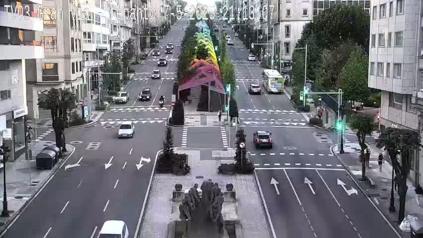 A city intersection with several cars and a pedestrian crossing, featuring a colorful structure in the center, under overcast skies.