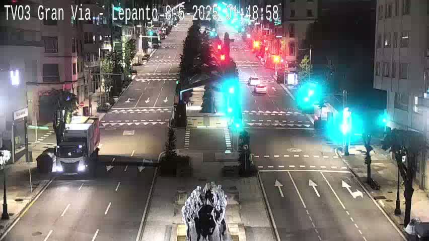 A nighttime street scene shows a mostly empty roadway with a few vehicles, traffic signals, and a statue in the center of the intersection.