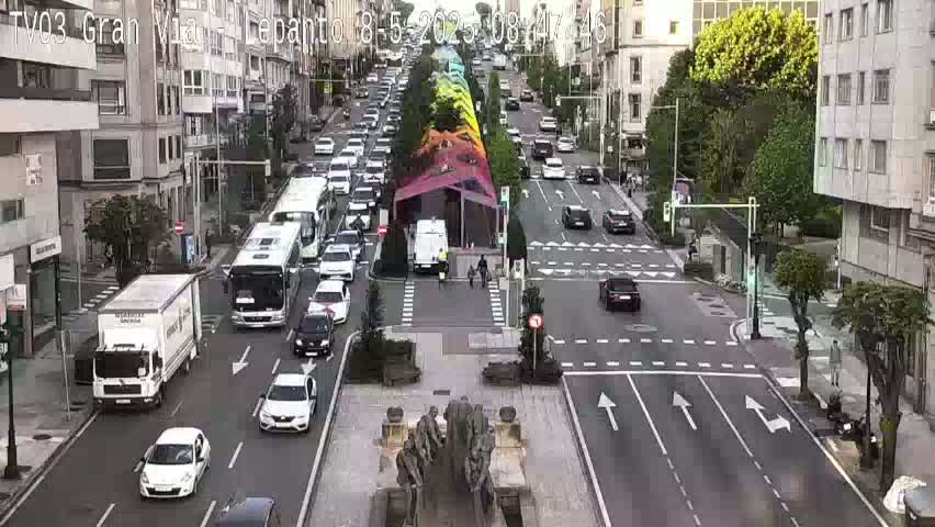 A city street scene shows multiple cars and trucks in motion on a sunny day, with a colorful structure in the center of the street and a large statue in a plaza.