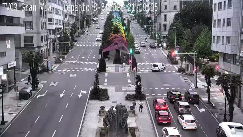 A city street scene on an overcast day shows several cars at an intersection with a large sculpture in the center of the road.