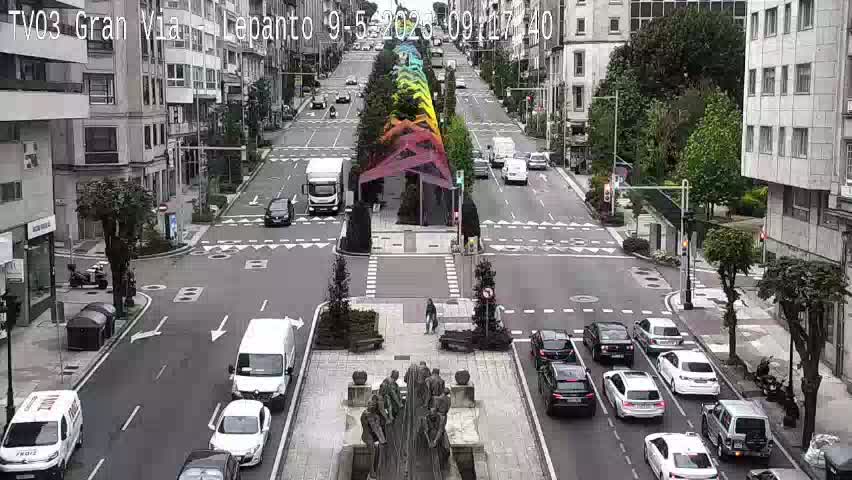 A city street scene on a partly cloudy day shows several cars stopped at an intersection, with a multi-colored structure in the middle of the street and a statue in a plaza.