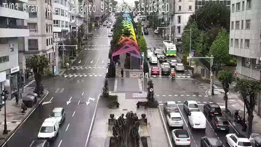 A rainy city street scene shows a wide intersection with multiple cars and a statue in the center, surrounded by buildings and a rainbow-colored structure.
