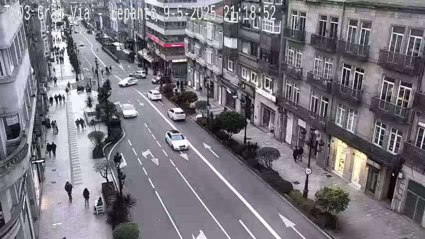A city street scene on an overcast day shows pedestrians walking on a sidewalk next to a multi-lane roadway with several cars driving in both directions, flanked by multi-story buildings.