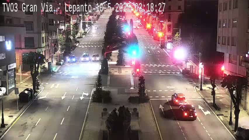 A nighttime, city street scene shows several vehicles at an intersection with traffic lights, and buildings lining the street.