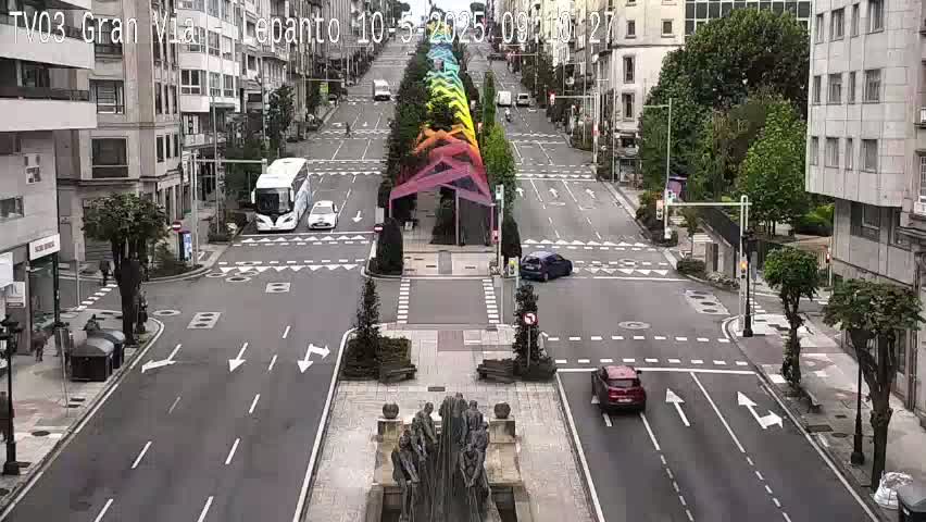 A wide, mostly empty city street with a few cars, a bus, and a rainbow-colored sculpture in the center, under a partly cloudy sky.