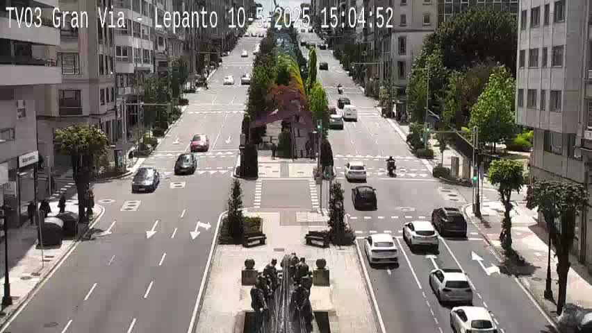 A sunny day on a city street shows several cars driving in both directions, with pedestrians near the buildings and a central monument featuring a sculpture.