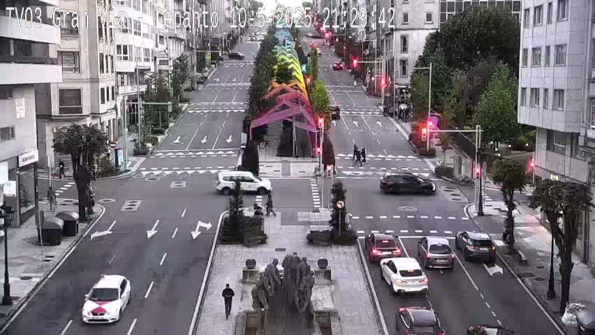 A city street scene with overcast weather shows cars driving past a central monument and a rainbow-colored structure.