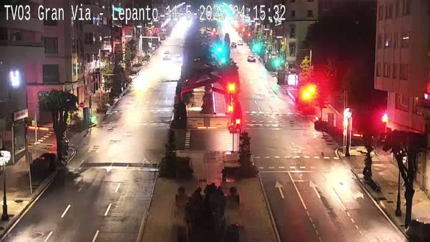 A wet, nighttime street scene shows a mostly empty multi-lane road with traffic lights and buildings lining both sides.