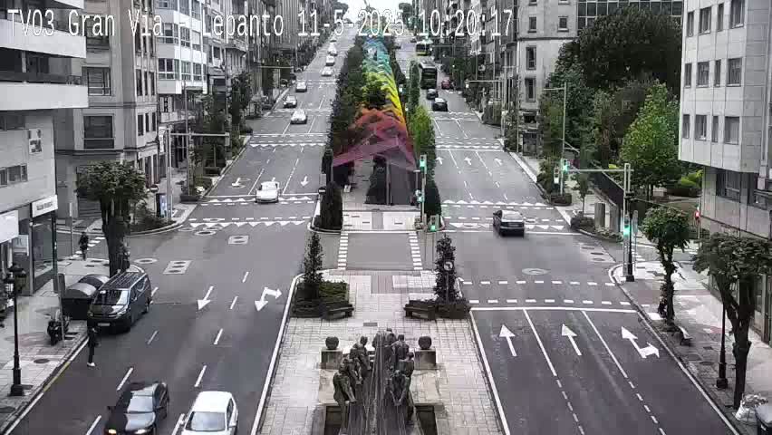 A wide, multi-lane street with several cars, a central median with a statue and colorful vertical structures, and buildings lining both sides on an overcast day.