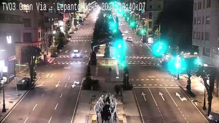 A nighttime, high-angle view of a mostly empty city street with traffic lights and a statue in the foreground.