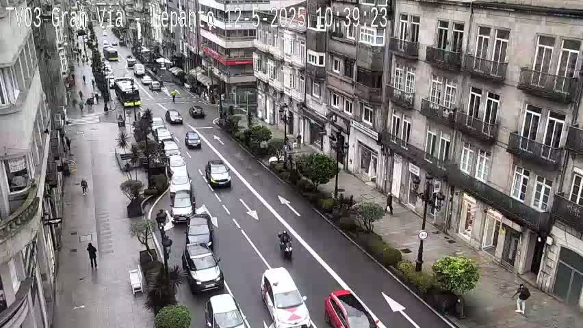 A city street scene on a wet day shows several cars driving in both directions, pedestrians walking on the sidewalk, and buildings lining the street.