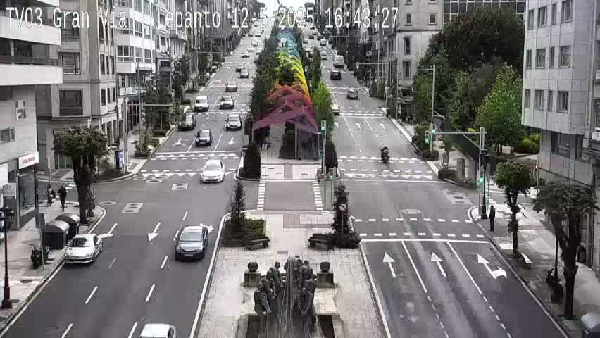 A city street scene shows several cars driving past a large fountain and multi-colored canopy structure under an overcast sky.