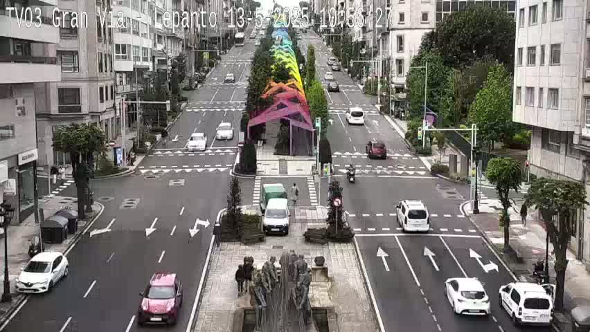 A city street scene on an overcast day shows cars driving on a multi-lane road, with a rainbow-colored structure in the center and a sculpture in the foreground.