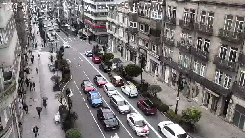 A city street scene on a partly cloudy day shows numerous cars driving down a multi-lane road lined with buildings and pedestrians walking on sidewalks.