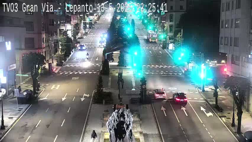 A nighttime, city street scene shows several cars driving on a multi-lane road that intersects with another road, with traffic lights, and a statue in the center of the intersection.