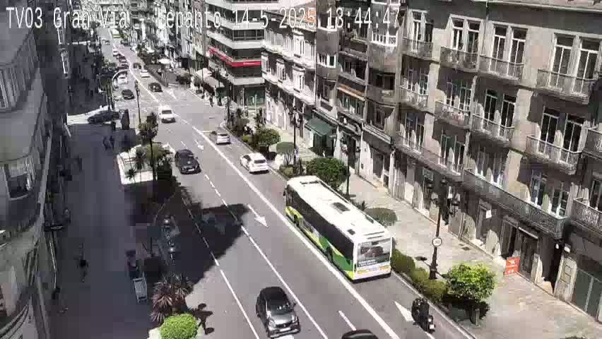 A city street scene on a sunny day shows several cars and a bus traveling down a multi-lane road lined with buildings.