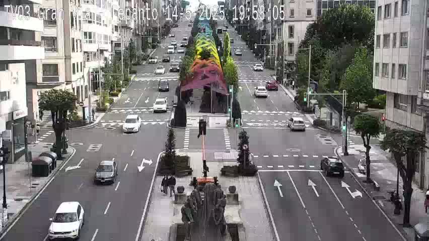 A city street scene shows vehicles moving along a roadway, with a large, colorful, abstract sculpture at a central intersection under a sunny sky.