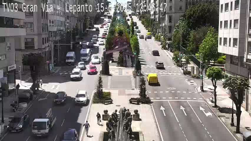 A sunny city street scene shows several cars driving on a multi-lane road that intersects with a cross street, with pedestrians walking near a central monument and trees lining the road.