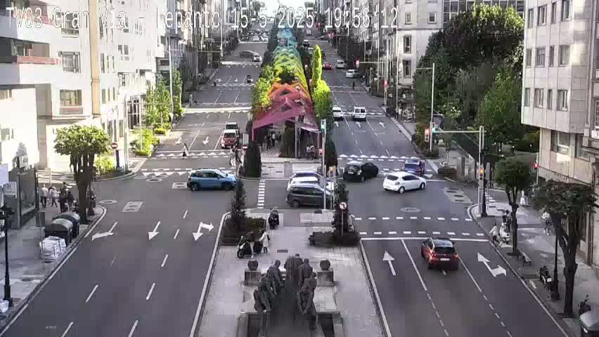 A city street scene shows several cars at an intersection under a partly sunny sky, with a colorful canopy structure in the center of the road and a statue at the intersection.