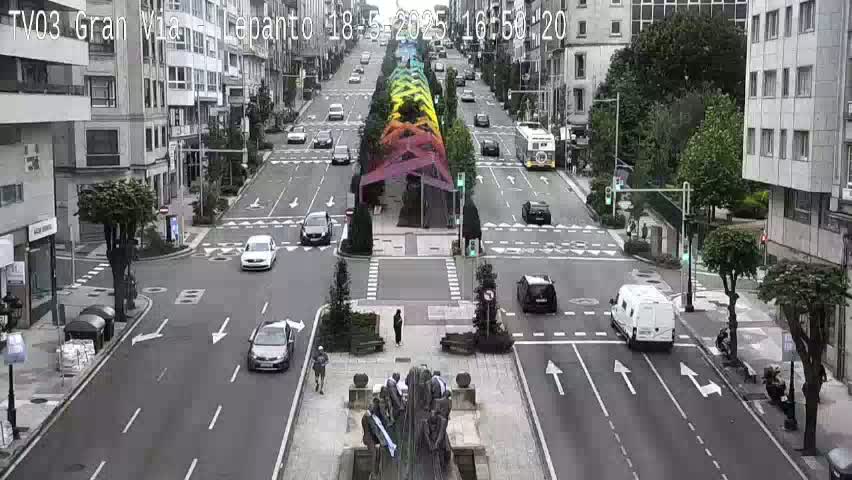 A city street scene on an overcast day shows several cars driving on a multi-lane road that intersects with another road, with a rainbow-colored structure in the center of the main road.
