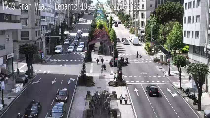 A sunny city intersection shows several cars navigating a multi-lane street, with pedestrians walking along the sidewalks and a central plaza featuring a sculpture.