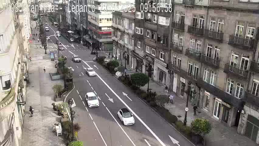 A city street scene on a partly cloudy day shows several white cars driving on a two-lane road that runs between tall buildings with balconies.