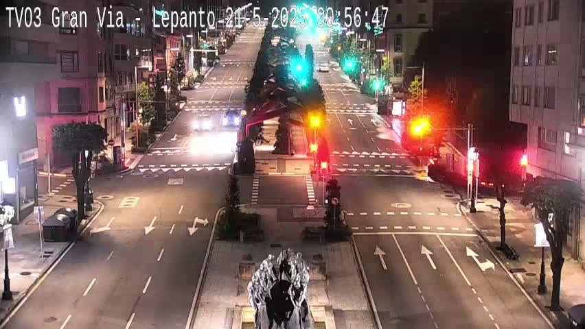 A nighttime, city street scene shows a sparsely trafficked intersection with several buildings lining the road, and a monument in the center of the intersection.