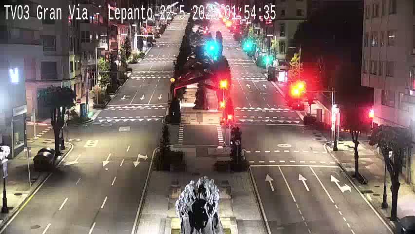 A mostly empty city street at night, illuminated by streetlights, shows traffic signals and a monument in the center.