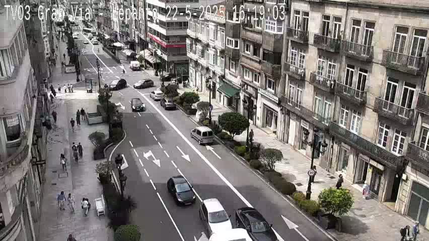A city street scene on a partly sunny day shows several cars driving on a multi-lane road that runs past multi-story buildings with pedestrians walking on sidewalks.
