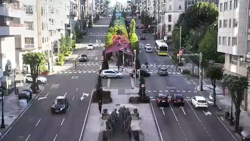 A city street intersection with several cars and a bus, under a partly sunny sky, features a central plaza with a large statue.