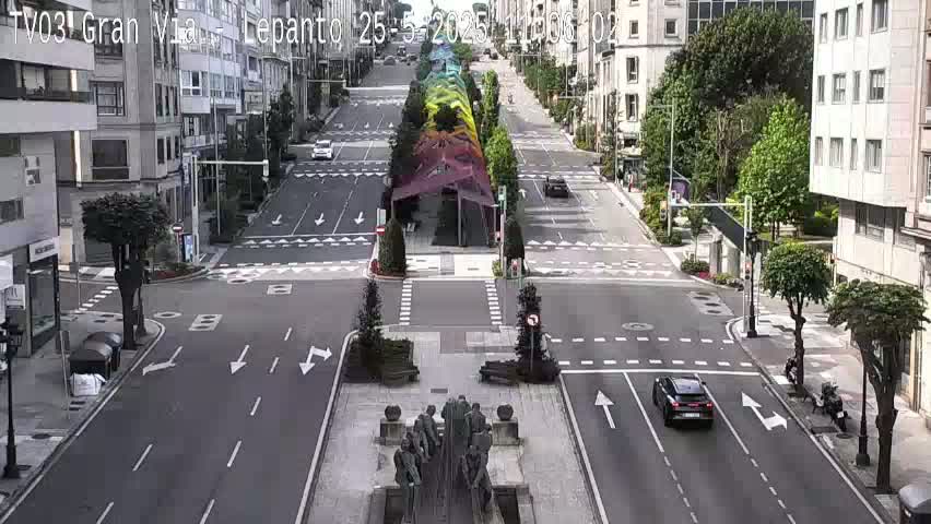 A wide, city street with few cars, lined with buildings and trees, features a central, colorful, arched canopy structure and a statue at an intersection on a partly sunny day.