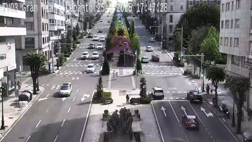 A sunny city street scene shows several cars at a crossroads near a pedestrian crossing and a statue, with buildings lining the street on either side.