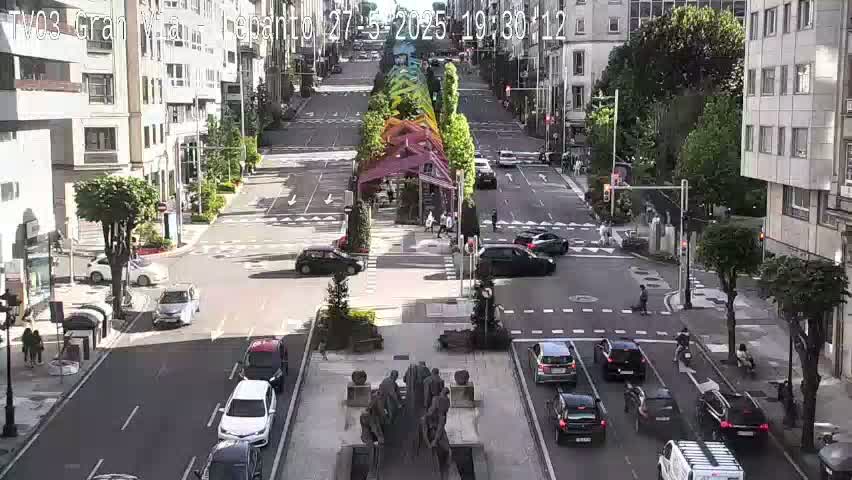 A sunny city street scene shows a wide intersection with multiple vehicles and pedestrians, centered around a large, dark-colored statue.