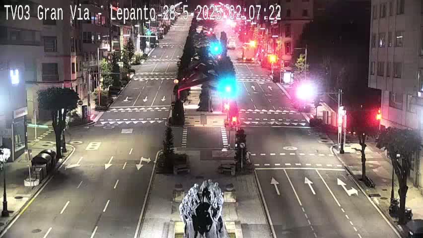 A nighttime, high-angle view of a mostly empty city street intersection with traffic lights and a statue in the center.