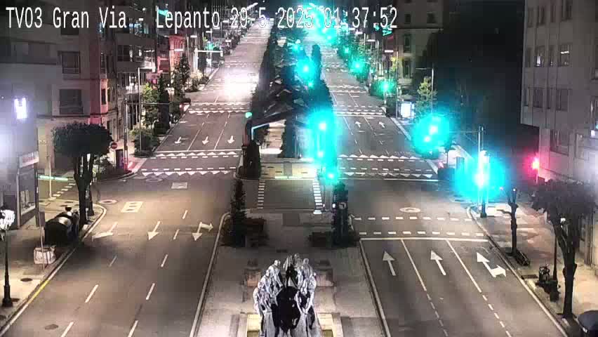A mostly empty, multi-lane road at night, with traffic lights and buildings lining either side, under clear skies.