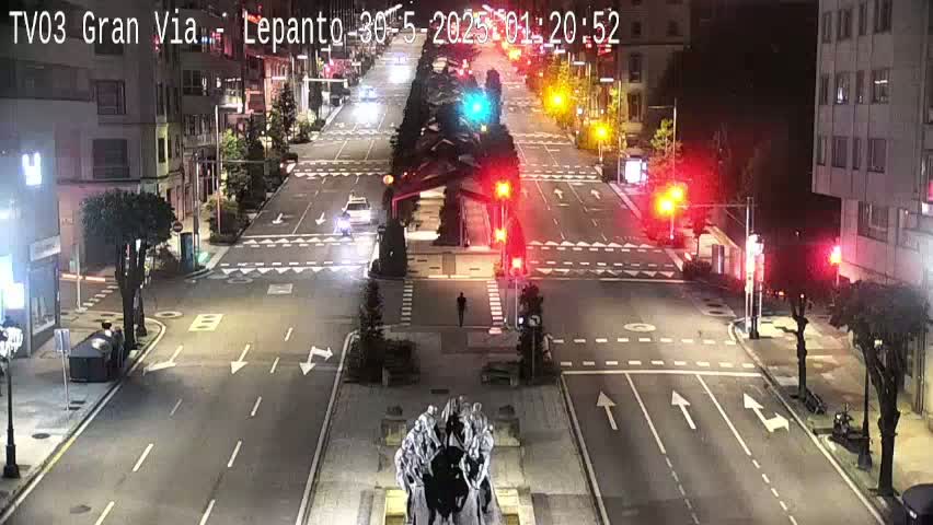 A nighttime street scene shows a mostly empty intersection with traffic lights and a statue in the center, under clear skies.