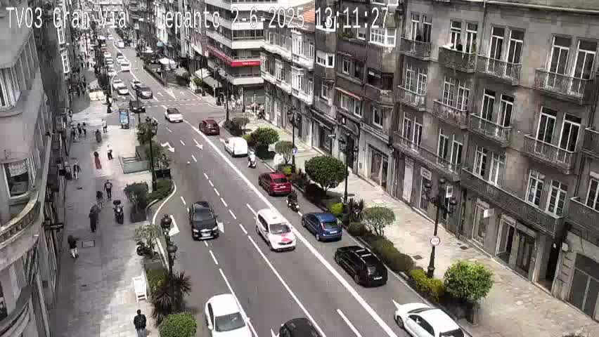 A city street scene on a sunny day shows several cars and pedestrians moving along a wide street lined with multi-story buildings.