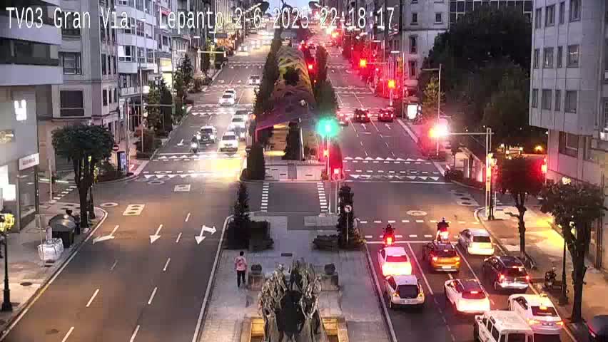 A city street scene at night shows cars driving past buildings and a traffic intersection under partly cloudy skies.