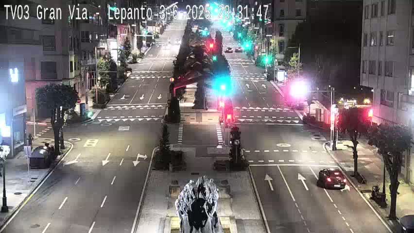 A nighttime view of a city intersection with several cars and traffic lights, under clear skies.