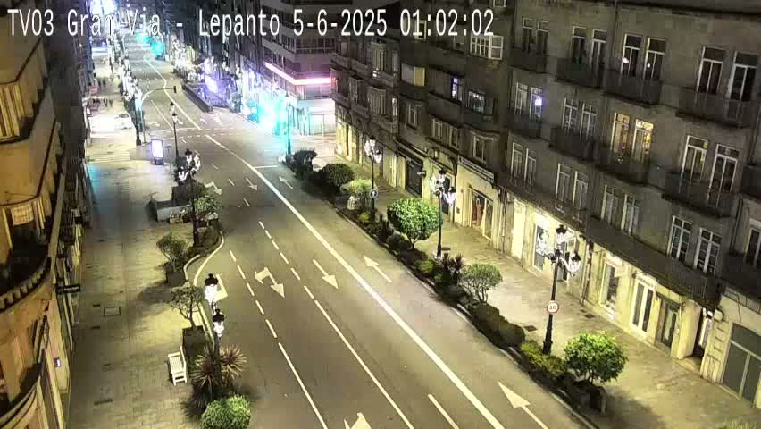 A nighttime, high-angle view of a mostly empty city street lined with buildings, featuring streetlights and sparse landscaping.