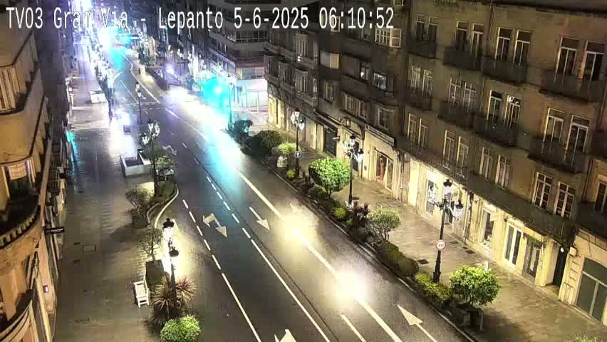 A wet, nighttime street scene shows a two-lane road with sparse traffic flanked by multi-story buildings with streetlights illuminating the sidewalks and landscaping.