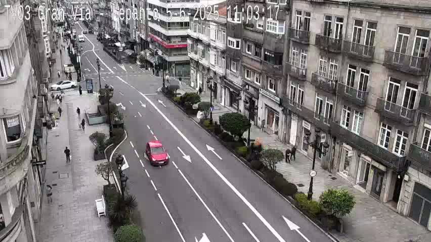 A mostly empty city street lined with multi-story buildings under an overcast sky, with a few pedestrians and a single red car visible.