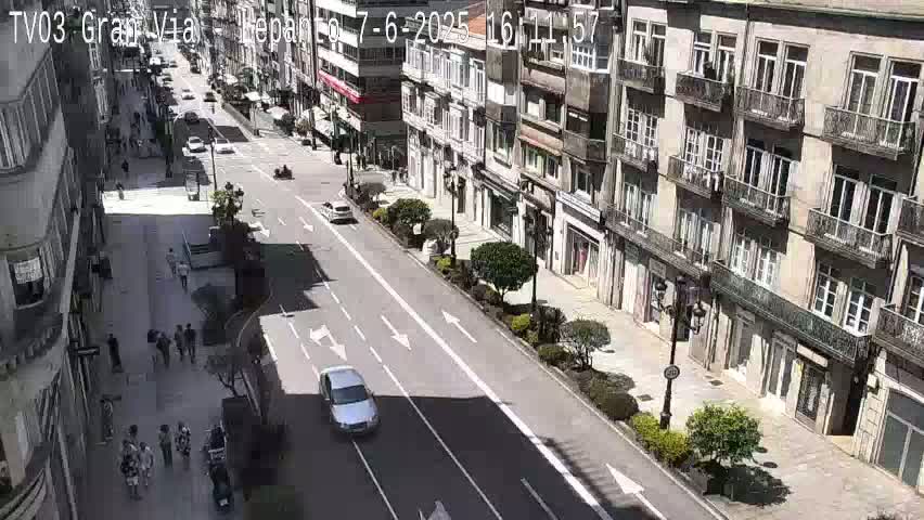 A sunny day on a city street shows several cars driving down a multi-lane road flanked by pedestrians on sidewalks and tall buildings with balconies.