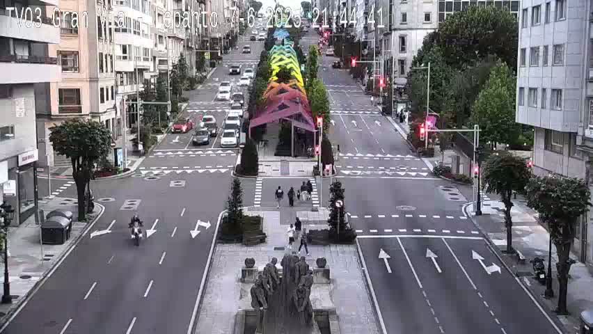 A city street scene with light traffic under overcast skies features a rainbow-colored structure in the center of the road.
