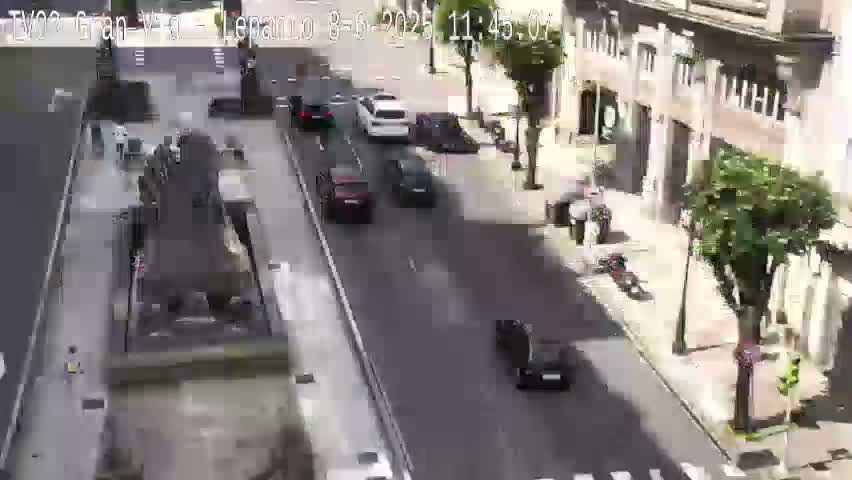 A sunny city street scene shows several cars driving on a road next to buildings, with a large fountain in the foreground.
