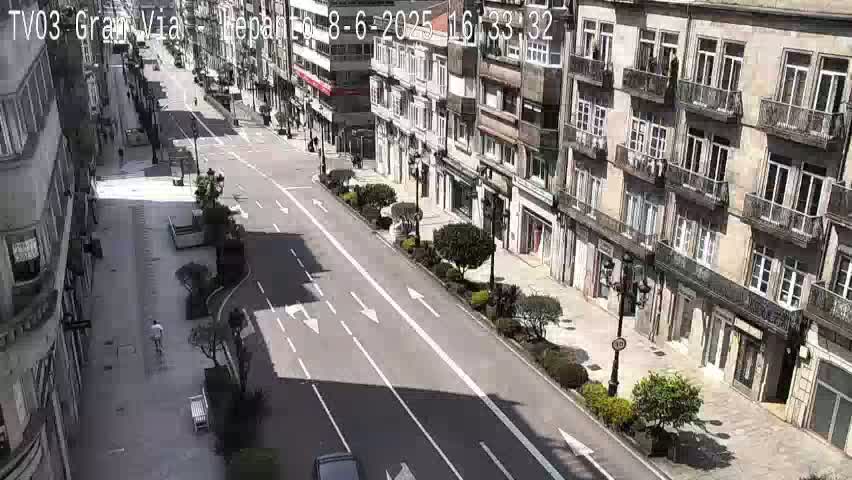 A sunny city street lined with multi-story buildings, featuring a pedestrian walking on the sidewalk and cars parked along the road.