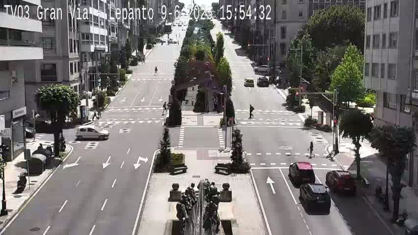A sunny day on a city street with several cars driving and pedestrians walking, a central median with landscaping and a sculpture, and tall buildings lining the street.