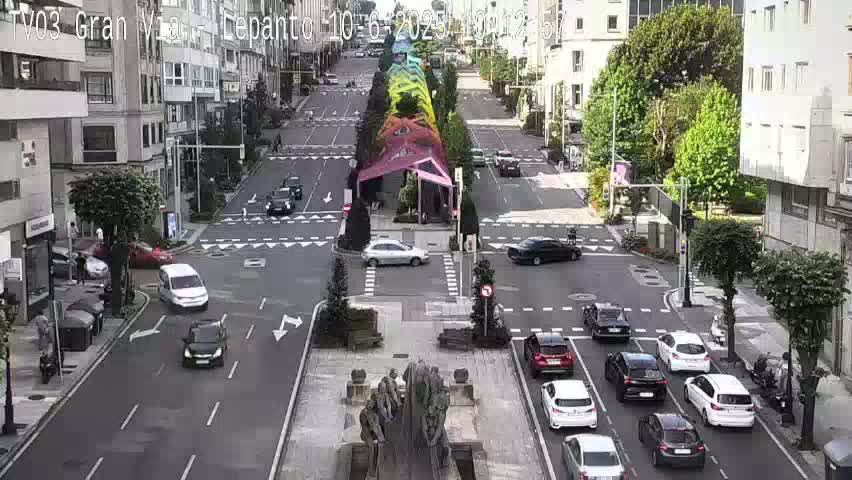 A city street scene with light traffic, featuring a central plaza with a statue and a colorful canopy, under a partly sunny sky.